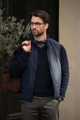 A man wearing a navy unisex vest over a sweater, carrying a brown bag, stands outdoors near plants.