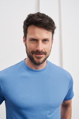 A blue t-shirt modelled by a man with short hair and a beard, featuring a logo on the shirt.