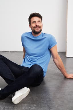 A blue T-shirt displayed on a seated male model wearing dark jeans and white sneakers, with a relaxed expression.