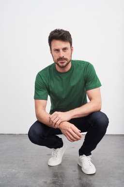 A green T-shirt worn by a man squatting in jeans and white sneakers, featuring a logo.