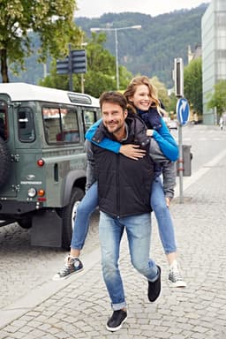 A padded men's vest in black, worn by a man giving a piggyback ride to a girl, with a city backdrop.