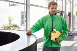 A man in a bright green softshell jacket stands near a black counter, holding a yellow folder and a notepad.