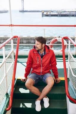 A man in a red maritime jacket sits on green stairs by the sea, wearing shorts and white shoes. The jacket has a logo.