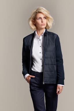 A ladies' hybrid sweat jacket in navy, featuring a zip front, a logo, and a stylish fit paired with a button-up shirt.