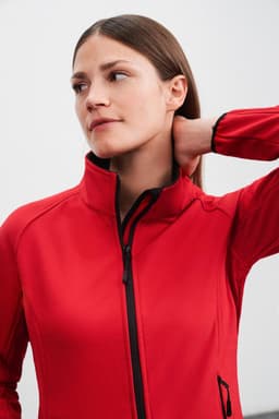 A red ladies softshell jacket with a high collar and a zip front, featuring a logo on the side.