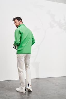 A man in a green softshell jacket stands against a white wall, wearing light trousers and holding a ball.