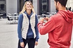 A smiling woman wears a white down vest over a blue hoodie, while a man in a red hoodie makes a heart shape with his hands.
