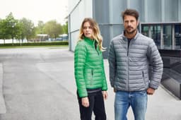 A green puffer jacket worn by a woman and a grey puffer jacket worn by a man stand outside a modern building.
