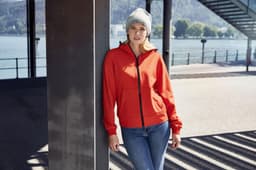 A red ladies hooded softshell jacket worn by a woman leaning against a post, paired with a beanie and jeans.
