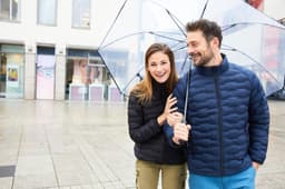 A modern padded jacket in navy, worn by a smiling man and woman under a clear umbrella in a city setting.