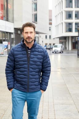 A man wearing a navy padded jacket stands on a city street. He has a beard and is dressed in blue trousers.
