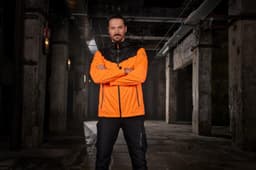 An orange and black men's allweather jacket is displayed on a model in an industrial setting. The jacket has a logo.