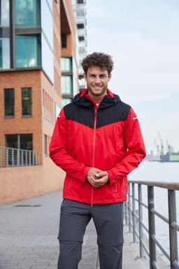A man wearing a red and black all-weather jacket, standing near a waterfront with buildings in the background.