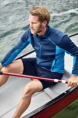 A man in a blue zip-up top and navy shorts sits in a rowing boat, holding a red paddle against a water backdrop.