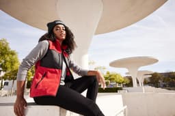 A woman is wearing a red and black fleece vest with a grey long-sleeve top, seated outdoors against a modern backdrop.