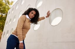 A knitted jumper in tan with a white collar, worn by a woman against a textured wall with circular cutouts.