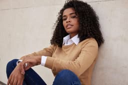A knitted round-neck pullover in tan, featuring a woman sitting against a wall, wearing a collared shirt underneath.