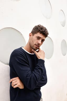 A model in a navy round-neck pullover stands against a textured wall with circular cutouts. The sweater features a white collar.