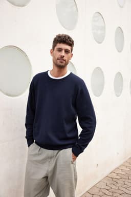 A man wearing a navy round-neck pullover with a white collar, posing in front of a textured white wall.