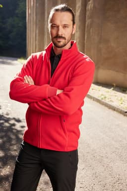 A man wearing a red bonded fleece jacket stands with arms crossed outdoors. The background features a stone wall.