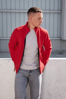 A man in a red bonded fleece jacket and grey t-shirt stands against a minimalist background. The jacket features a logo.
