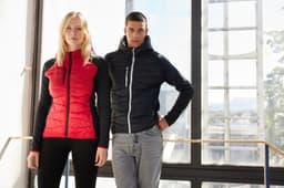 A red and black hybrid jacket worn by a woman and a man, featuring a logo and set against a bright indoor background.