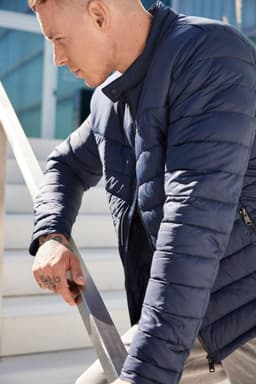 A navy padded jacket with a zip front and a high collar, styled on a man leaning against a railing.