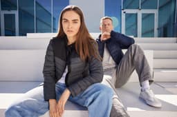 A padded jacket in navy blue, worn by a seated model and another model in the background, both against a modern architecture backdrop.