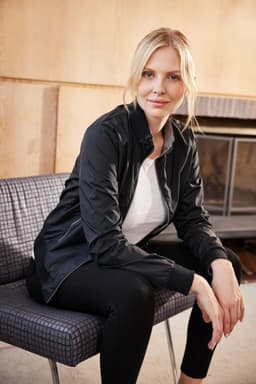 A black ladies' blouson jacket displayed on a model sitting on a chair, with a fireplace in the background.
