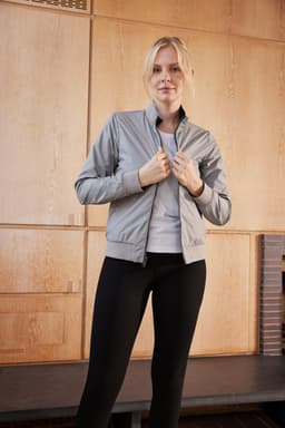 A light grey ladies blouson jacket is worn by a model, paired with black leggings against a wooden backdrop.