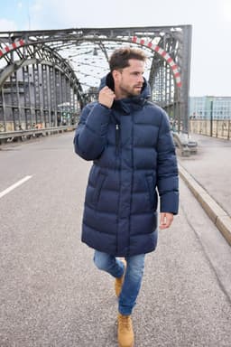 A man walks on a bridge wearing a navy long puffer coat, paired with jeans and tan boots.