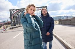 A puffer jacket in dark blue and a long coat in teal, both worn by people on a city street.