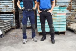 Two individuals wearing workwear pants in navy and black, standing against a textured background of metal containers.