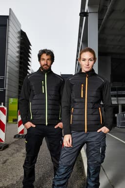 A black hybrid jacket for ladies featuring orange accents, worn by a model in an industrial setting.