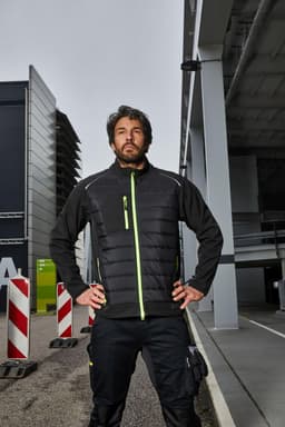 A hybrid jacket in black with neon green accents, featuring a zip front and a logo, worn by a model outdoors.