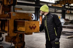 A men's hybrid jacket in black with bright yellow accents, worn by a man in a beanie and gloves near industrial equipment.