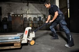 A man in dark workwear pants pulls a pallet jack in a warehouse setting. The pants feature pockets and a logo.