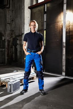 Workwear pants in blue and black, worn by a person standing in an industrial setting. The outfit includes a black polo shirt.