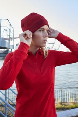 A woman wearing a red long-sleeve polo shirt and a red beanie, standing by the water. The scene features a harbour backdrop.