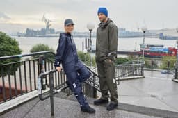 Two models wearing waterproof rain pants in navy and olive green, standing on an outdoor staircase with a waterfront backdrop.