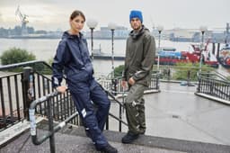 A navy blue and olive green rain jacket worn by two models, standing near a waterfront with ships in the background.