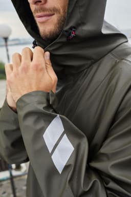 A model wearing an olive green rain jacket with a hood, featuring a logo on the sleeve.