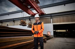A man in an orange workwear jacket stands outside a warehouse, wearing gloves and a hard hat. The setting includes steel beams and a crane.