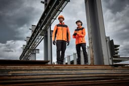 Two individuals wearing bright orange work jackets stand on metal beams at a construction site, with overcast skies in the background.
