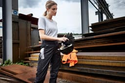 A woman in grey workwear-pants stands holding a helmet, with steel sheets in the background. The pants are dark in colour.