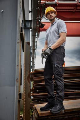 A man in a grey shirt and black work pants stands in front of construction materials, wearing a yellow hard hat and gloves.