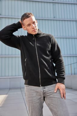 A black Sherpa Jacket with a zip front and side pockets, worn by a person standing against a modern background.