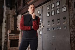 A woman in a black and red padded hybrid jacket stands thoughtfully beside a vintage control panel.