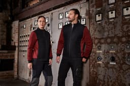 A pair of models wear a ladies padded hybrid jacket in black and red against an industrial backdrop. The jacket features branding.
