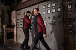 A padded hybrid jacket in black and red, worn by a man and a woman in an industrial setting. The jackets feature branding.